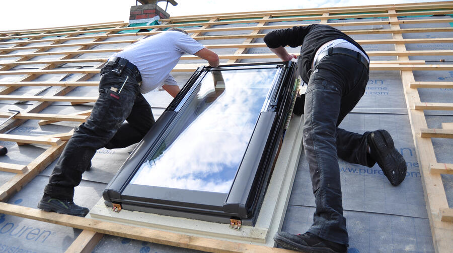 Two workers installing a glass skylight on a roof under construction, with wooden framing and a plastic weather barrier underneath.
